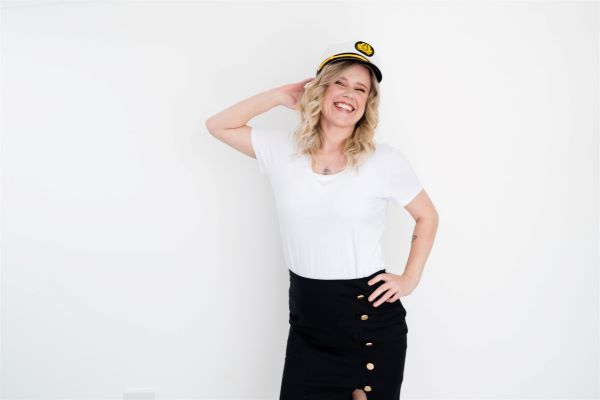 Chloe wearing a white top, one hand behind her head and the other on her hip. Wearing a ship captain's hat